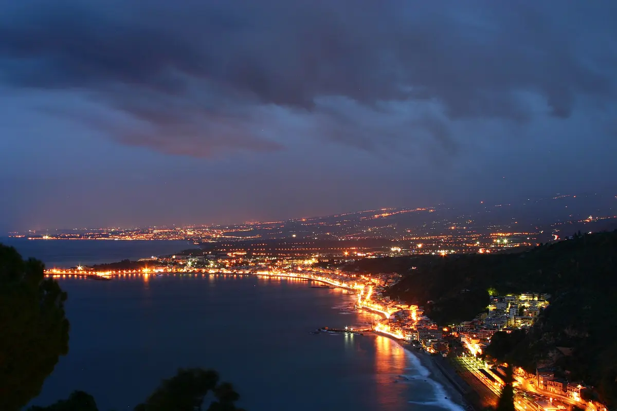 As luzes da costa em torno do Golfo de Naxos à noite, vistas de uma encosta de Taormina sob um céu escuro e nublado, com uma linha brilhante de estrada ao longo da costa.