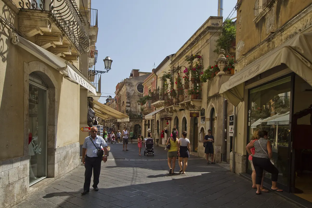 O Corso Umberto em Taormina de dia, uma rua pedonal estreita entre edifícios de pedra antiga com toldos de lojas e varandas de ferro, e algumas pessoas a caminhar.