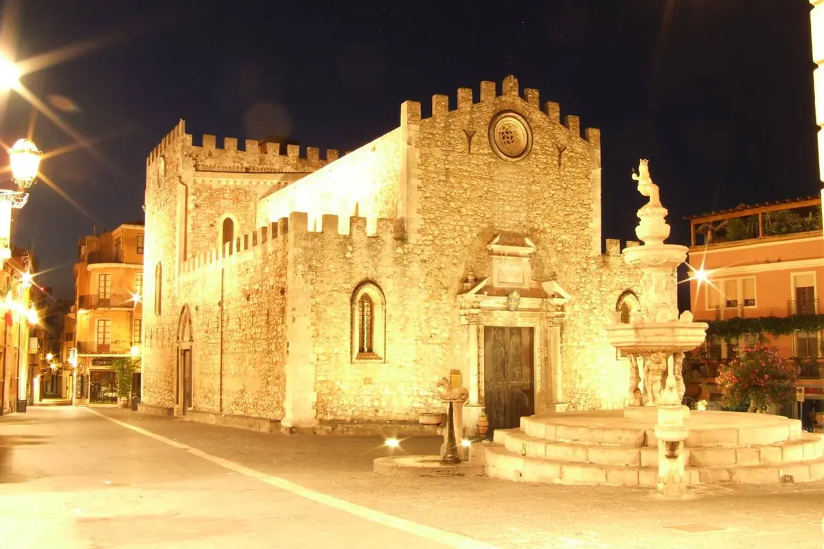 A fachada ameada da Catedral de Taormina iluminada a ouro à noite, com a fonte barroca da Piazza Duomo à sua direita.