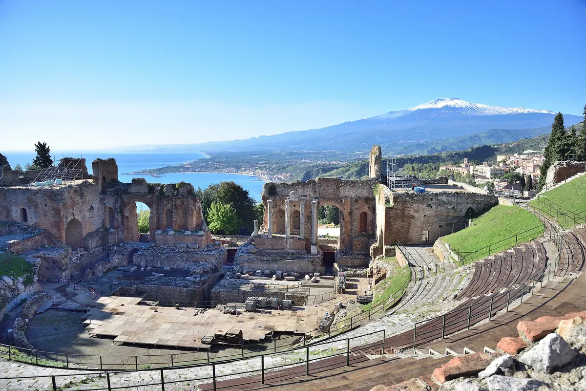 A parede cénica em ruínas e os assentos curvos do antigo teatro de Taormina num dia claro, com o cone nevado do Monte Etna e o mar ao longe.
