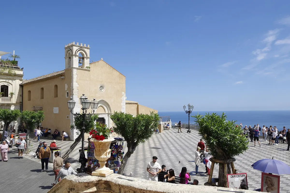 A Piazza IX Aprile em Taormina de dia, com o pavimento em xadrez de pedra, árvores em vasos, a igreja de San Giuseppe e uma faixa de mar azul para além do muro do terraço.
