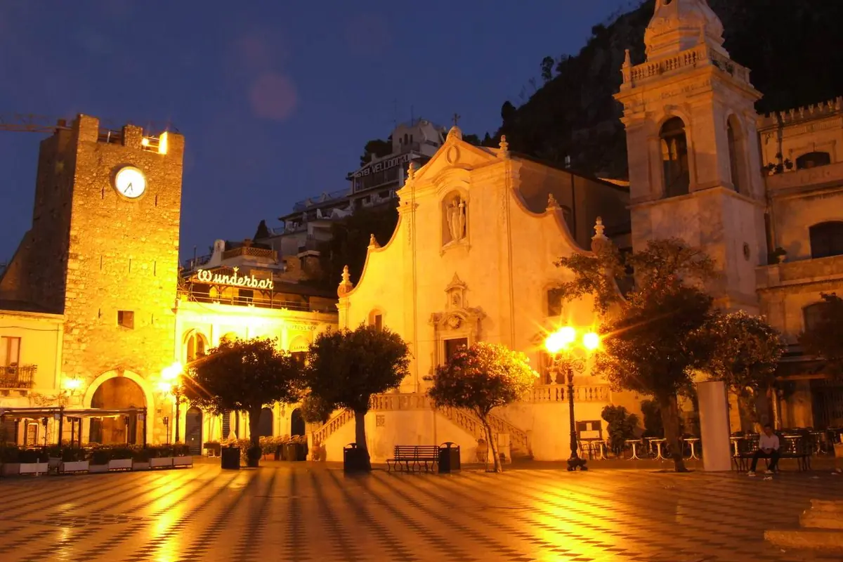 Piazza IX Aprile em Taormina à noite, com a torre do relógio iluminada à esquerda, a fachada dourada da igreja de San Giuseppe e um campanário à sua direita sobre uma praça axadrezada.