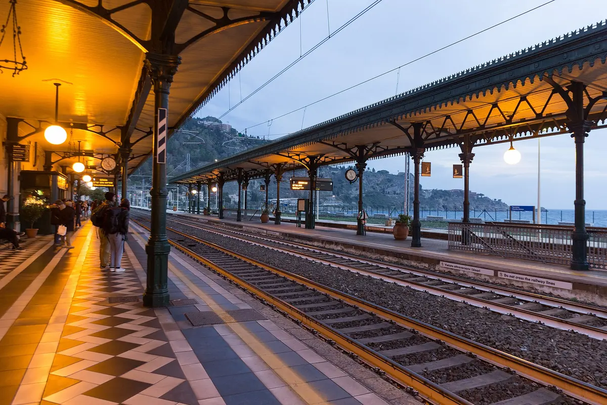As plataformas da estação de Taormina-Giardini sob uma longa cobertura de ferro, com um pavimento de ladrilho xadrez, candeeiros e as colinas acima da linha.