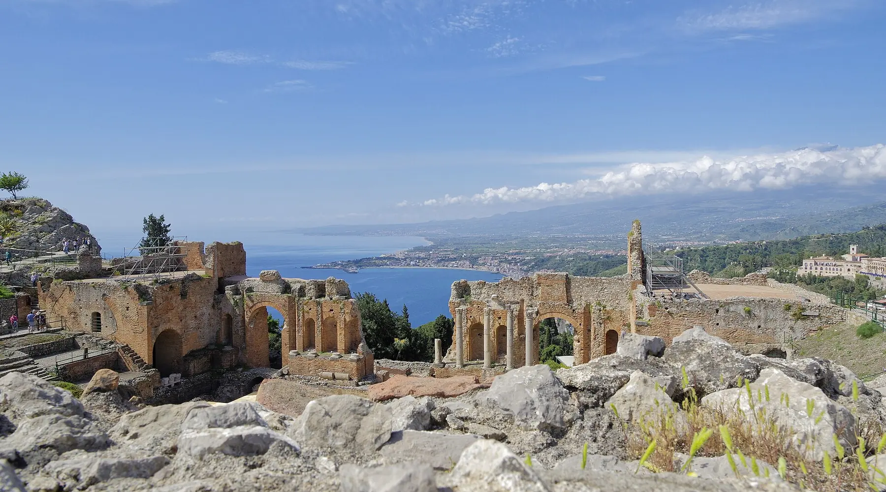 Os arcos em ruínas do antigo teatro de Taormina vistos através de rocha irregular, com a linha da costa e um longo banco de nuvens no horizonte.