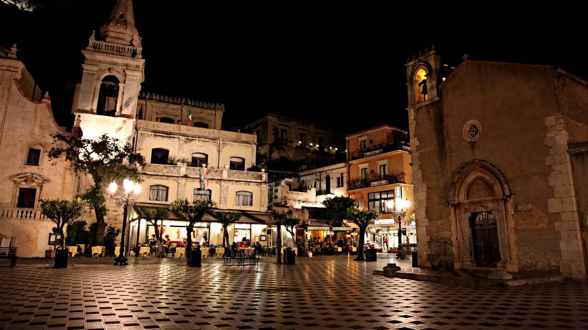 A Piazza IX Aprile em Taormina à noite, um pavimento de pedra xadrez iluminado por candeeiros de rua, com a torre sineira da igreja de San Giuseppe à esquerda e uma pequena igreja de pedra com um portal gótico à direita.